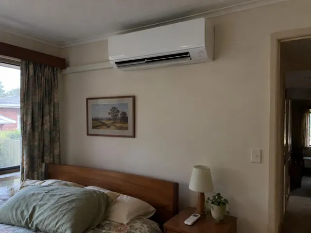 Clean split system mounted high on a bedroom wall above a bed with curtains and a bedside table in Elizabeth Grove